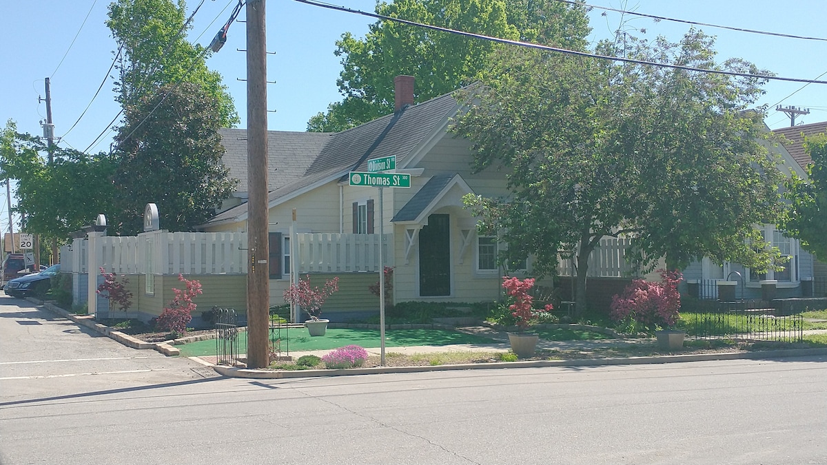 The exterior of a single-story building is showcased, featuring a welcoming entrance under a covered porch. Surrounding greenery, including trees and flowering shrubs, adds to the setting. A white picket fence provides a charming boundary, while street signs indicate the corner location.