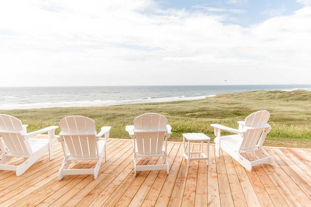 Four white Adirondack chairs are arranged on a wooden deck, facing a scenic view of the ocean. The calm sea stretches to the horizon under a partly cloudy sky, with lush green grass visible in the foreground.