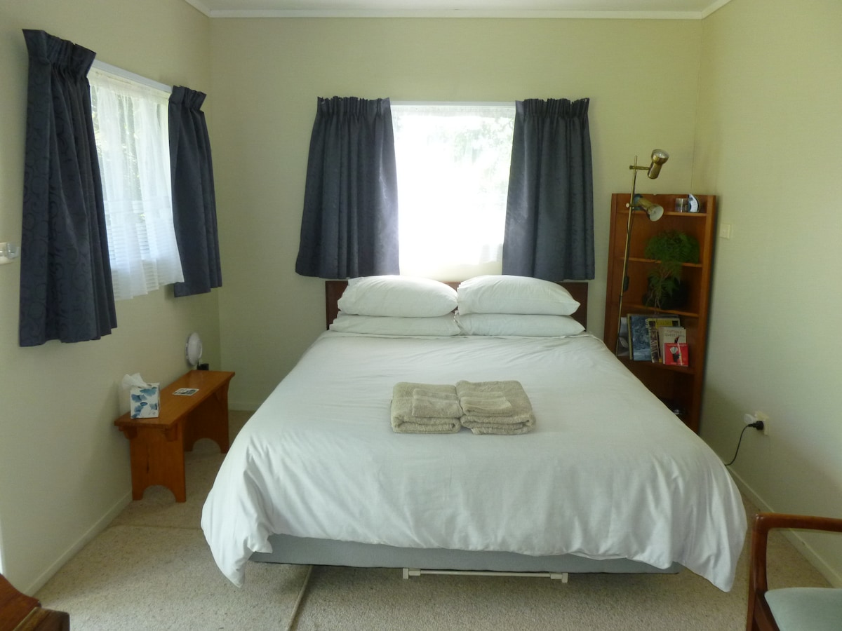 The bedroom features a spacious bed with neatly arranged pillows and a soft blanket. Natural light enters through a window, complemented by dark curtains. A bedside table stands beside the bed, and a small bookshelf is positioned nearby, showcasing personal items and decor.