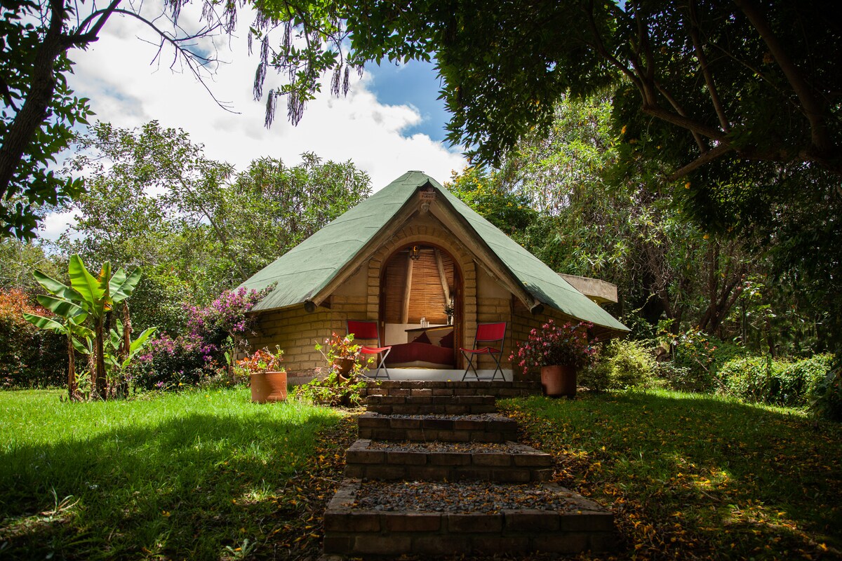 A charming cabin with a distinctive thatched roof is surrounded by lush greenery and flowering plants. A pathway of stone leads to the entrance, where two red chairs are positioned on a small patio, inviting relaxation in a tranquil outdoor setting.