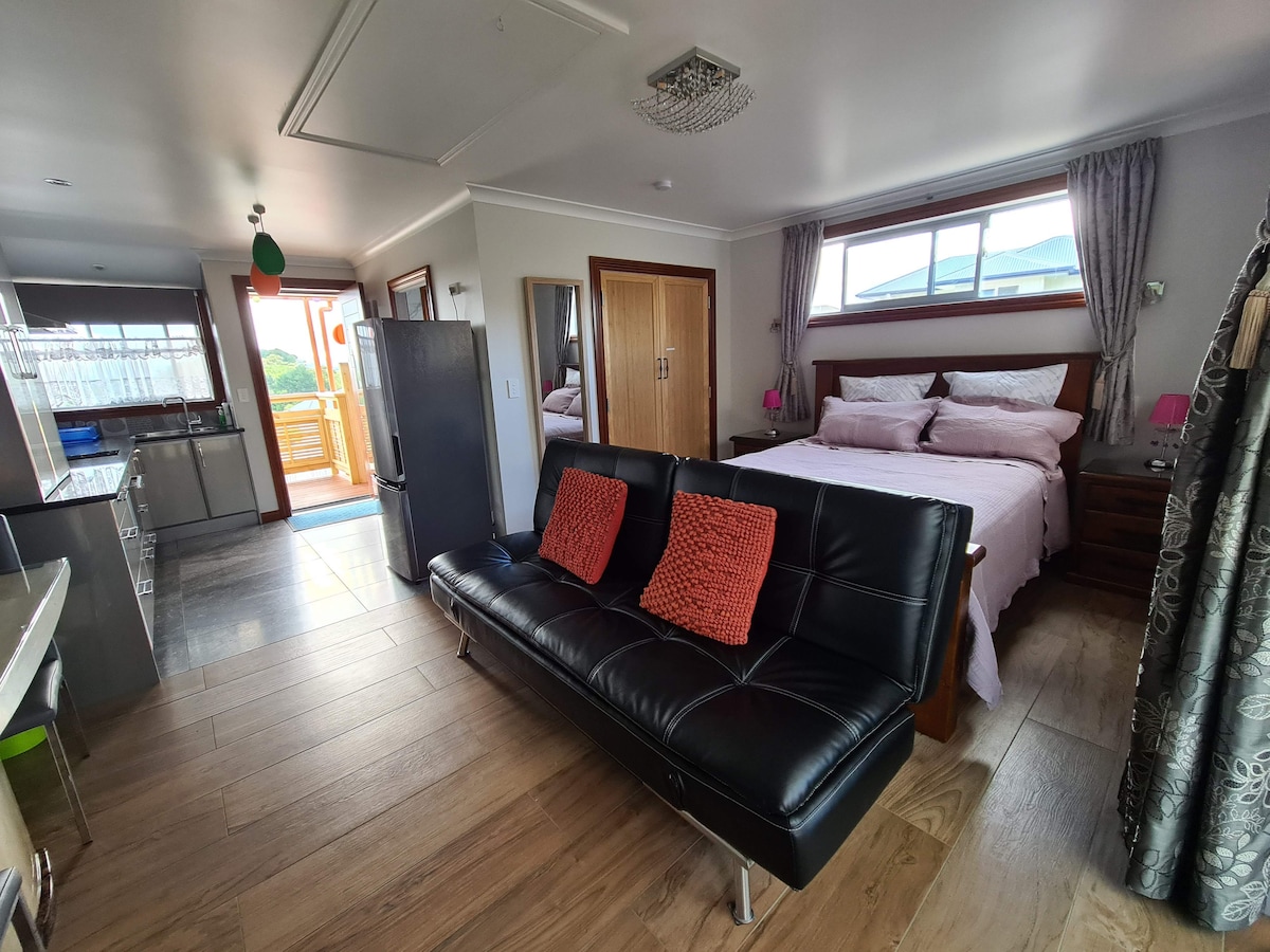 A modern bedroom is equipped with a spacious bed adorned with light pink linens. A comfortable black sofa with vibrant orange cushions is positioned in the living area. Natural light floods the space through large windows, creating a bright and airy ambiance. A glimpse of the outdoor deck is visible through the sliding door.