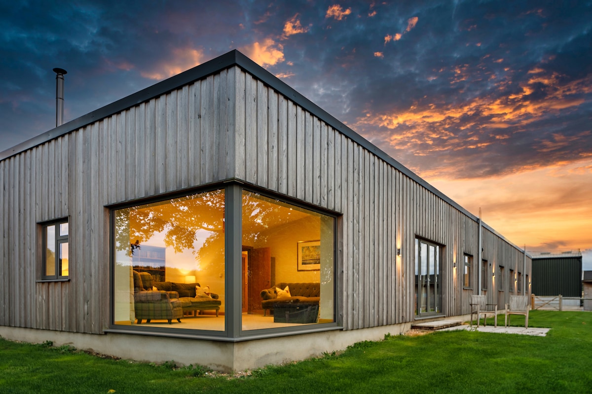 The exterior of The Pig House showcases modern wooden cladding against a backdrop of a vibrant sunset. Expansive glass windows reveal warm living spaces inside, while manicured grass surrounds the property, contributing to a welcoming environment.