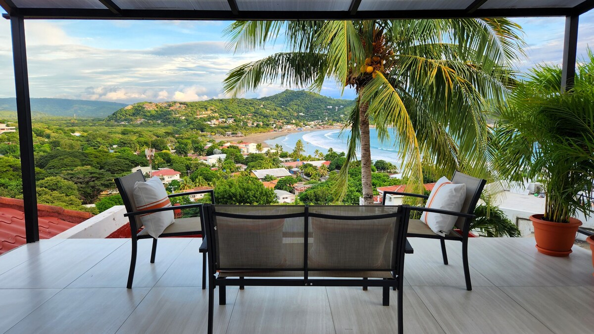 A spacious balcony features comfortable seating, offering sweeping views of the coastline and surrounding hills. Lush greenery and a palm tree frame the scene, while the ocean glistens in the distance under a clear sky.