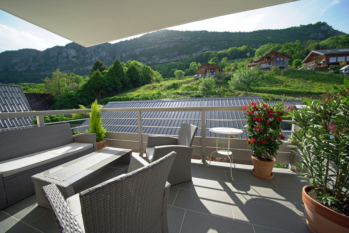 A serene balcony area features comfortable grey wicker furniture arranged around a low table. Lush greenery and blooming flowers are visible in pots, while a picturesque hillside with residential buildings adds a natural backdrop. The space is illuminated by natural light from the open sky.