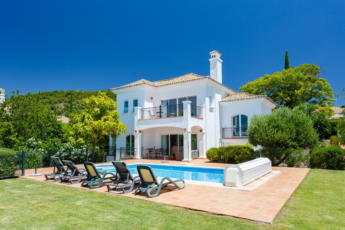 A two-story villa is presented with a private pool and sun loungers arranged on a spacious patio. The exterior features large windows and a balcony, surrounded by lush greenery and neatly trimmed lawns, under a clear blue sky.