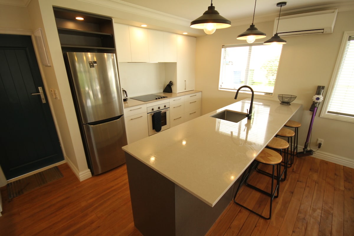 A modern kitchen is presented, featuring sleek white cabinetry and stainless steel appliances. A spacious island with a quartz countertop is paired with stylish bar stools. Natural light enters through a large window, enhancing the functional cooking space equipped with an oven, hob, and dishwasher.