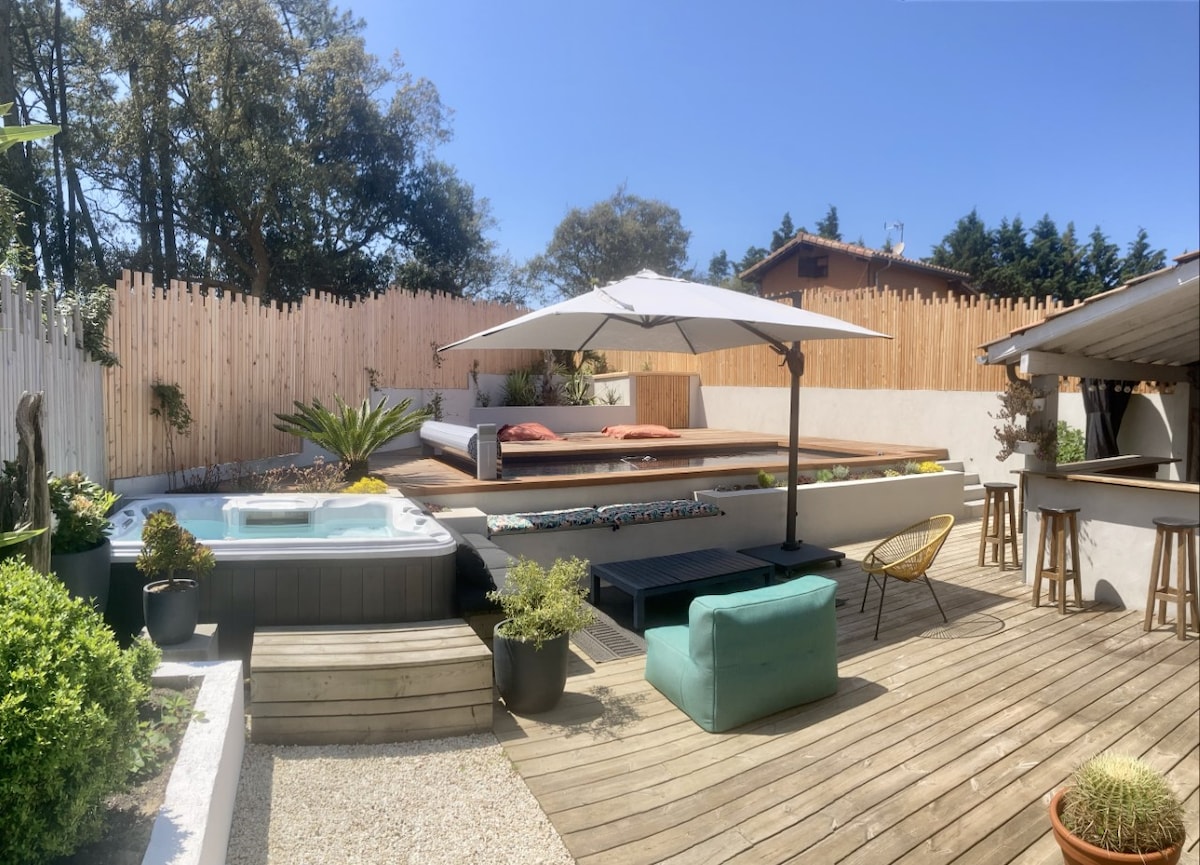 An outdoor space is presented featuring a spa and a wooden deck with lounge seating. An umbrella provides shade over a seating area, while a bar counter is visible in the background. Lush greenery surrounds the area, creating a relaxing environment for guests.