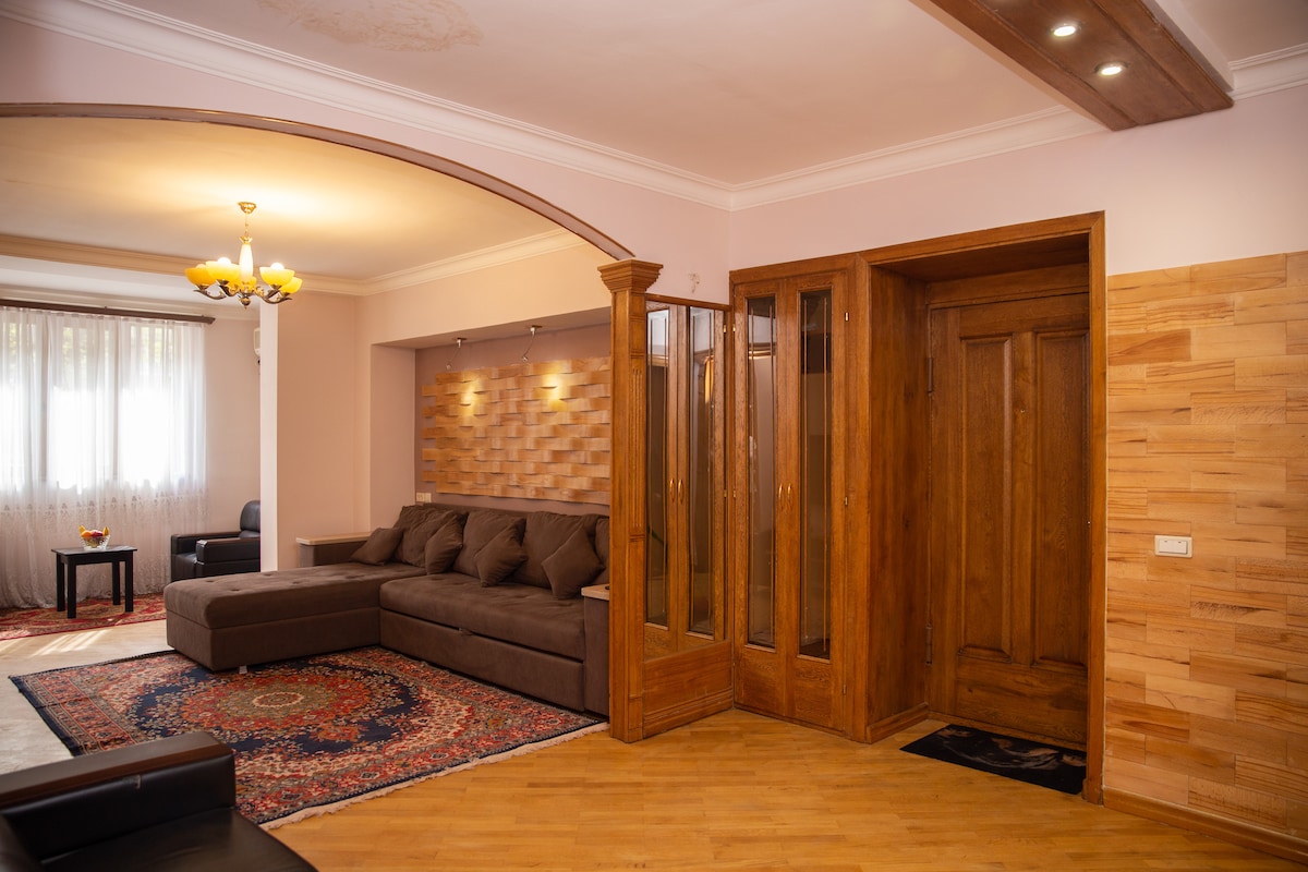 A spacious living area features a L-shaped brown sofa, invitingly arranged around a decorative rug. Natural light filters through sheer curtains, illuminating the cozy atmosphere. A wooden entrance with glass elements offers a welcoming transition into the room.