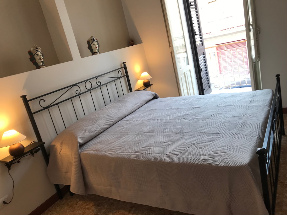 A cozy bedroom features a spacious bed made with a light-colored blanket. Soft bedside lamps provide gentle illumination. The room opens onto a balcony through large doors, allowing natural light to fill the space and offering views of the outside.