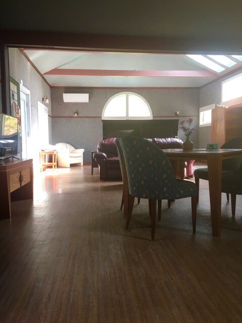 A spacious studio interior is illuminated by natural light streaming through large windows. Comfortable seating is provided by a dark leather couch and a light-colored armchair. A wooden dining table with chairs sits near the center, complementing the hardwood floor.