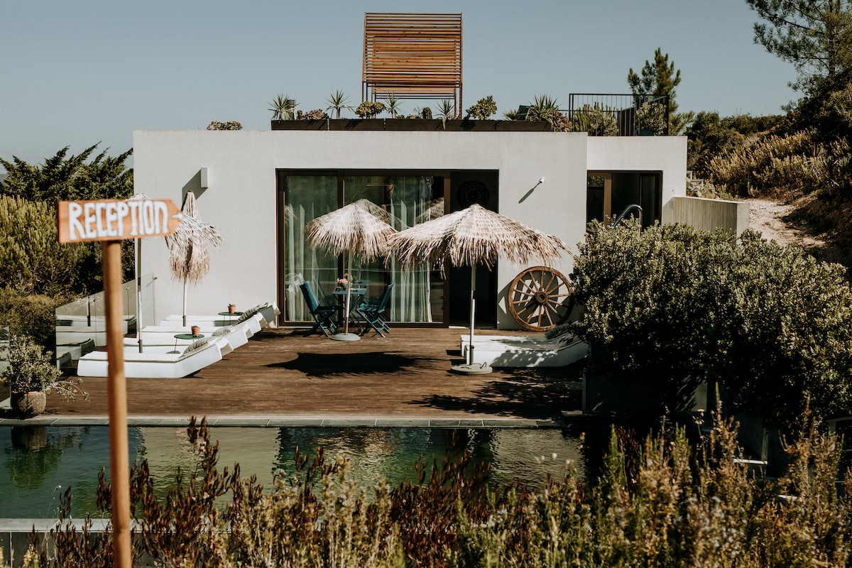 A modern eco-villa is presented, featuring a spacious deck with umbrellas providing shade. Lounge chairs are positioned next to the ecological pool, surrounded by lush greenery. A reception sign stands nearby, welcoming guests to this tranquil retreat.