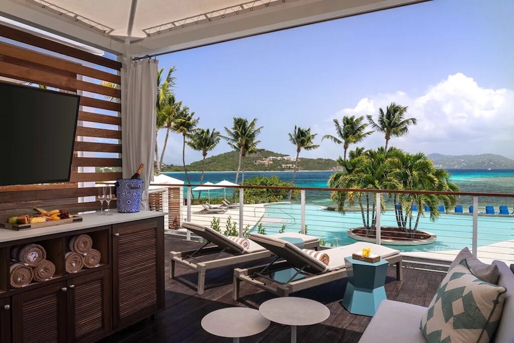 A spacious patio area is displayed, featuring two lounge chairs beside a low table. A partial railing offers a view of crystal blue waters and surrounding palm trees. A flat-screen television is mounted on wooden panels, enhancing the outdoor decor.