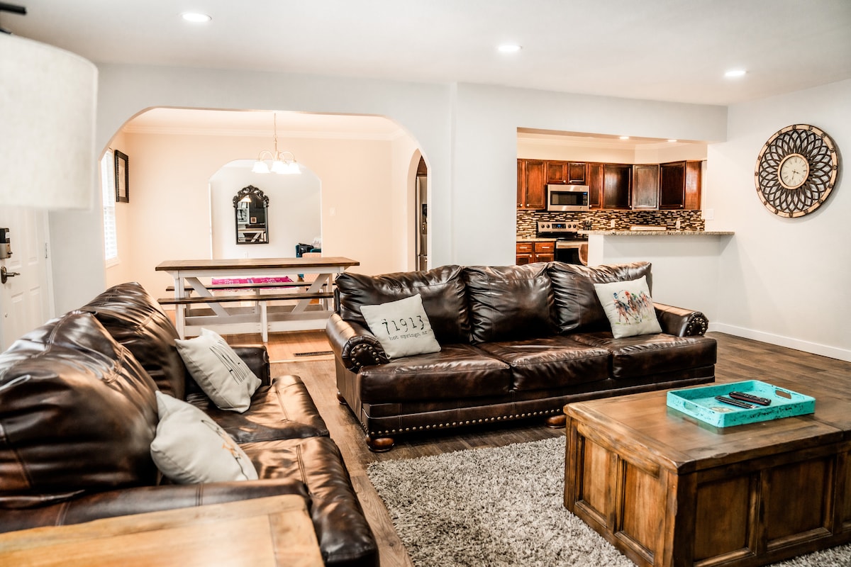 A spacious living area is highlighted by a large leather sectional sofa and a wooden coffee table. The room includes soft lighting and a decorative wall clock. An open dining area with a wooden table is visible in the background, leading into the kitchen.