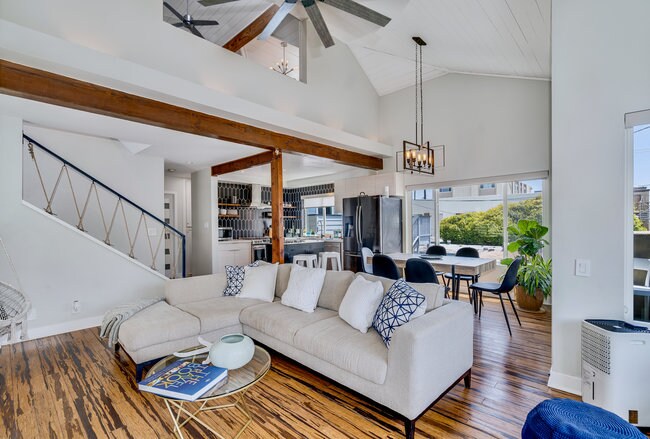 An airy living area is featured with high vaulted ceilings and a large sectional sofa adorned with blue and white pillows. A round coffee table with a decorative book is centered in the space, while a dining area with a table and chairs is visible through expansive windows.