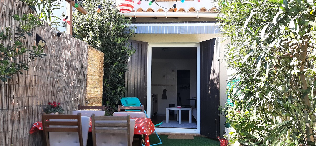 An outdoor courtyard is shown, featuring a set of wooden chairs around a table with a red tablecloth. Lush greenery and decorative lighting enhance the space, which is framed by wooden fences. Large doors lead into the living area, connecting indoor and outdoor environments.