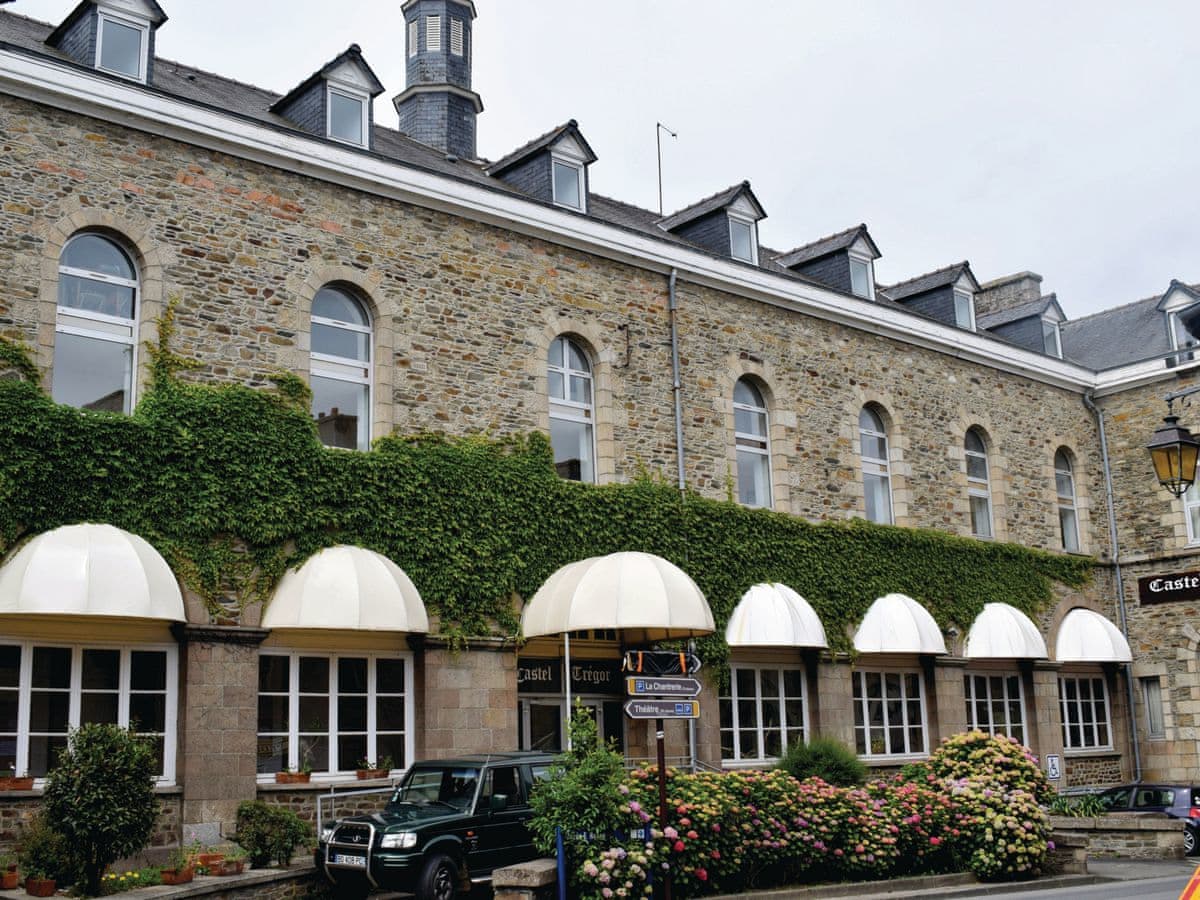 A historic building features a stone facade with multiple arched windows. White awnings provide shade to the lower-level entrances, while greenery climbs the exterior. Colorful flower beds add vibrancy to the entrance, and a parked vehicle is visible in front of the structure.