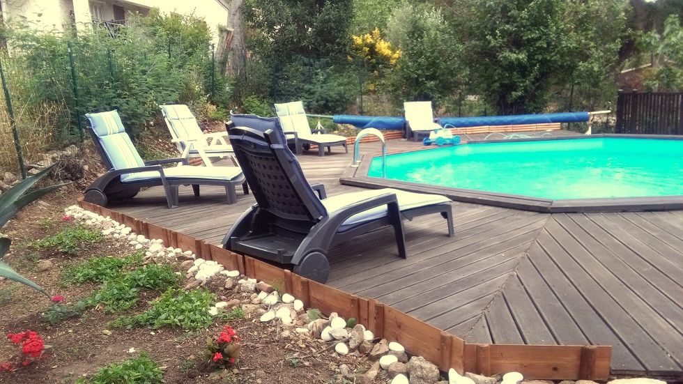 A private swimming pool is surrounded by a wooden deck, featuring several lounge chairs arranged for relaxation. Lush greenery and colorful flowers enhance the garden area, and a safety fence is visible around the pool, providing a secure environment.