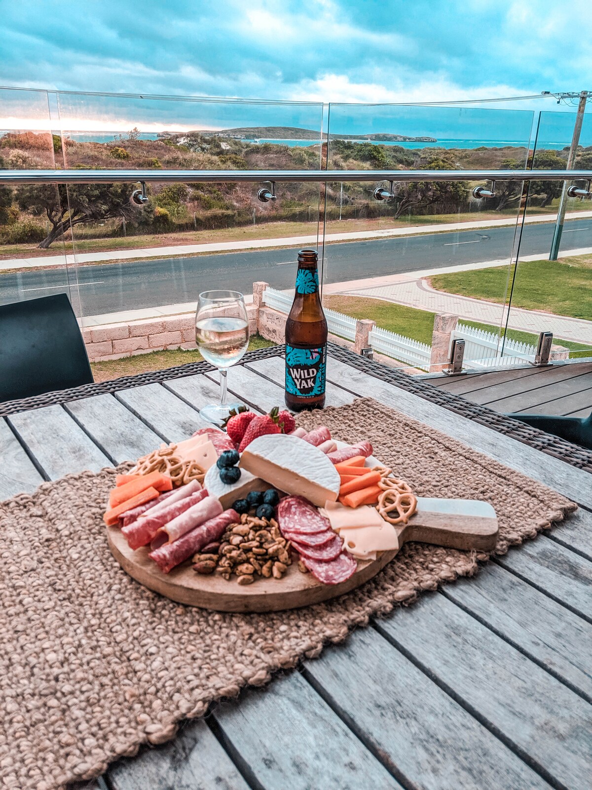 A wooden platter displays an assortment of cheeses, meats, nuts, and fresh fruit, set on a textured tablecloth. A glass of white wine and a beer bottle sit alongside the spread. A panoramic view of the outdoors is visible beyond the glass railing.