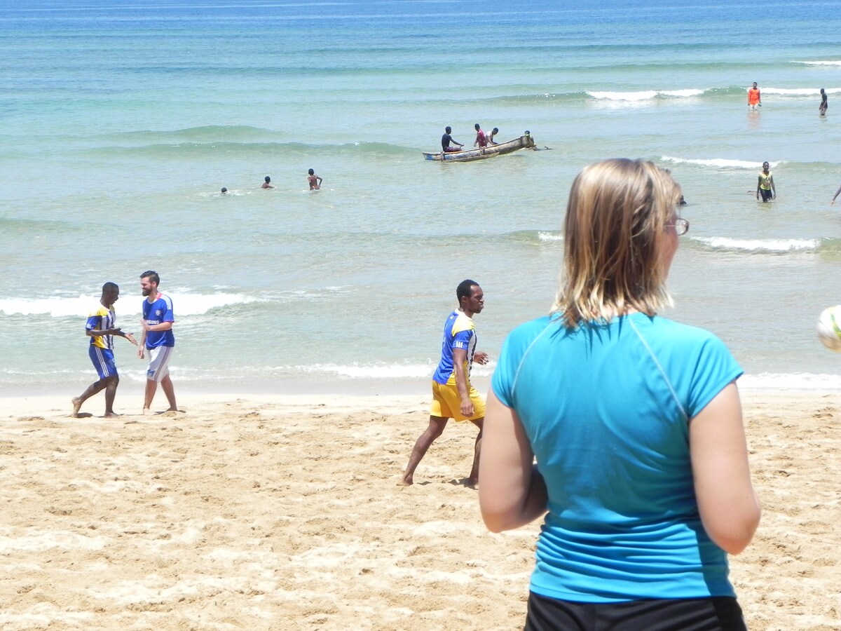 A sandy beach scene is portrayed, featuring individuals engaging in recreational activities near the shoreline. Gentle waves lap against the coast, while small fishing boats are visible in the distance. A person with shoulder-length hair stands in the foreground, observing the beachgoers.