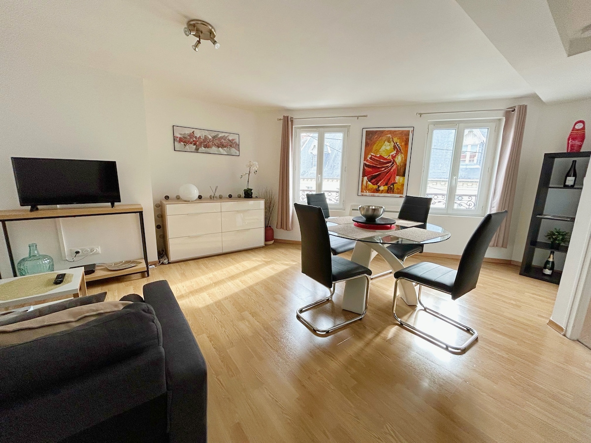 A bright living area features a round dining table surrounded by four chairs, with natural light streaming through three windows. A media console holds a television, while a tasteful painting adds color to the wall. The space is adorned with plants and decorative items.