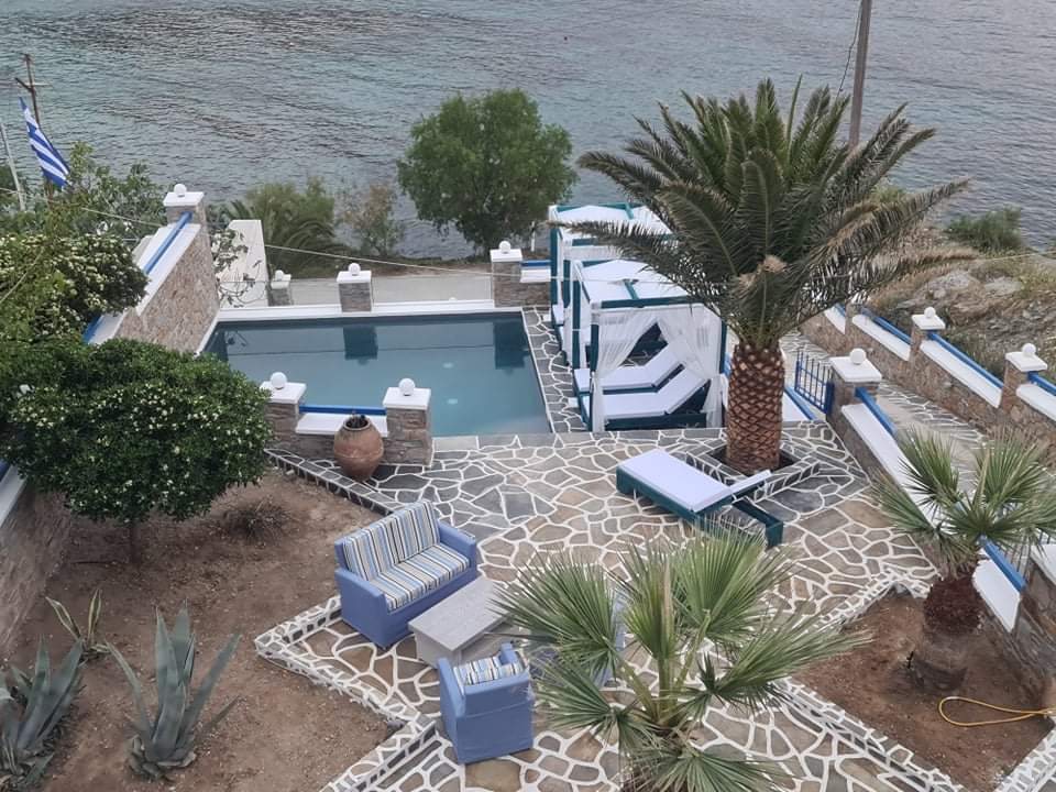 An outdoor area features a swimming pool surrounded by decorative stone pathways and lush greenery. Sun loungers are arranged beside the pool, and seating is provided in a cozy nook. Palm trees and other plants enhance the tranquil setting with views of the nearby water.