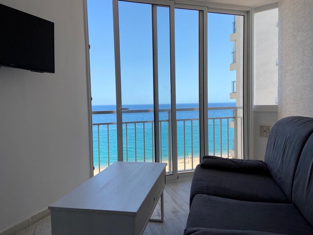 A living area features a dark sofa positioned in front of large glass doors, offering unobstructed sea views. A light-colored coffee table sits centrally, enhancing the open feel of the space. A wall-mounted television adds functionality and comfort to the room.