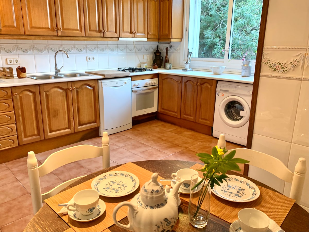 A functional kitchen is composed of wooden cabinetry and tiled walls. A round dining table is set with floral-patterned tableware, featuring a teapot and cups. A window allows natural light, and appliances including a dishwasher and washing machine are visible.
