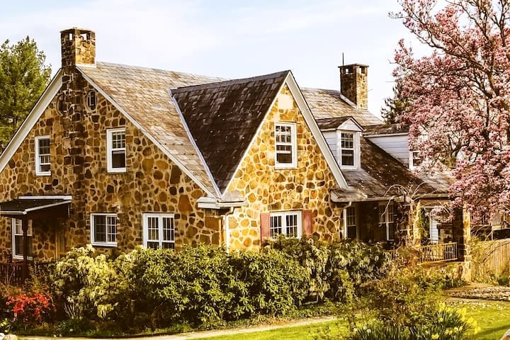 The historic stone exterior of the building is displayed, showcasing a charming design with a gabled roof and multiple chimneys. Lush greenery surrounds the structure, complemented by flowering trees, creating a serene atmosphere in a natural setting.