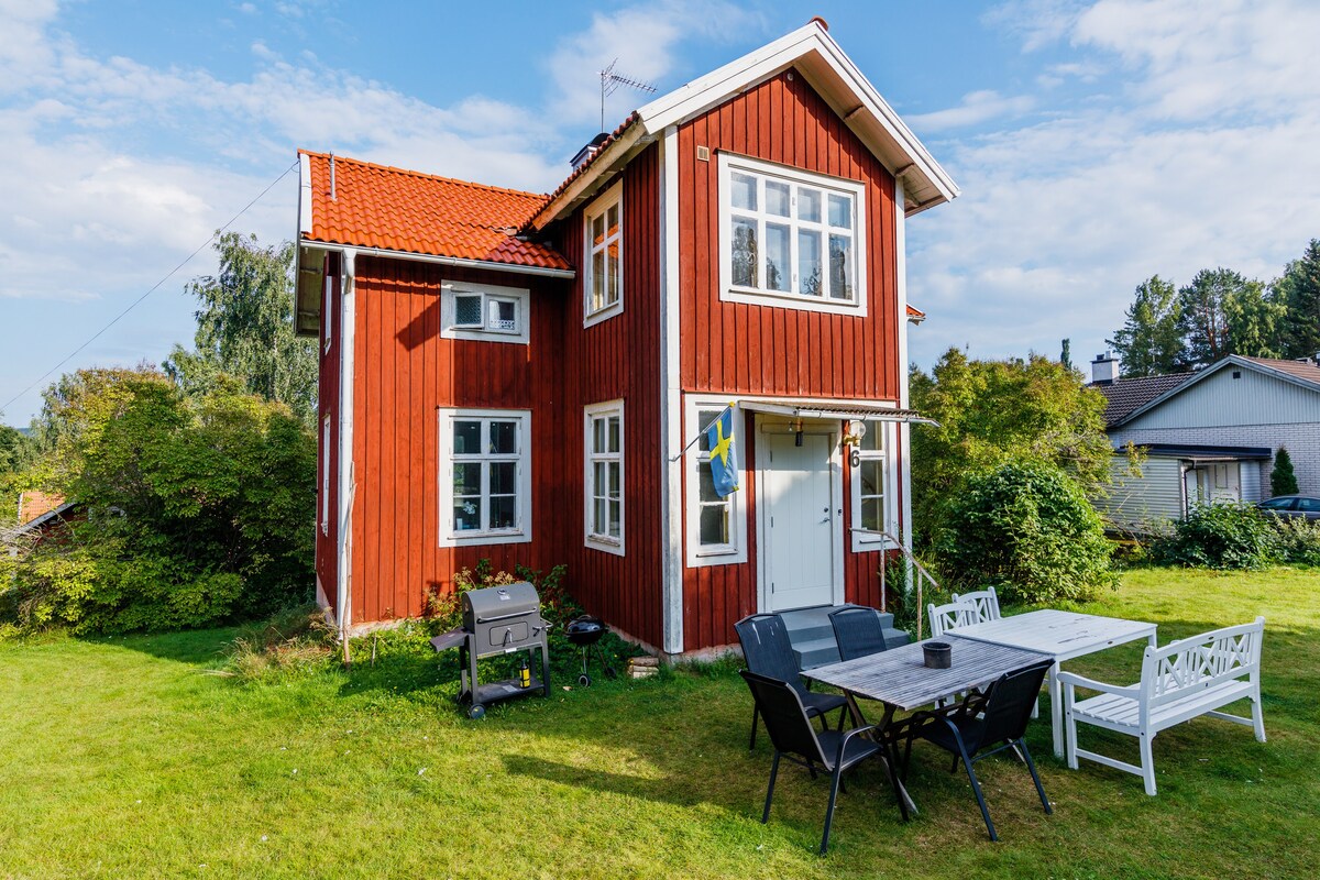 The charming red house features a traditional design with white trim and multiple windows. An outdoor seating area with a table and chairs is visible in front of the house, surrounded by lush greenery and open lawn space.