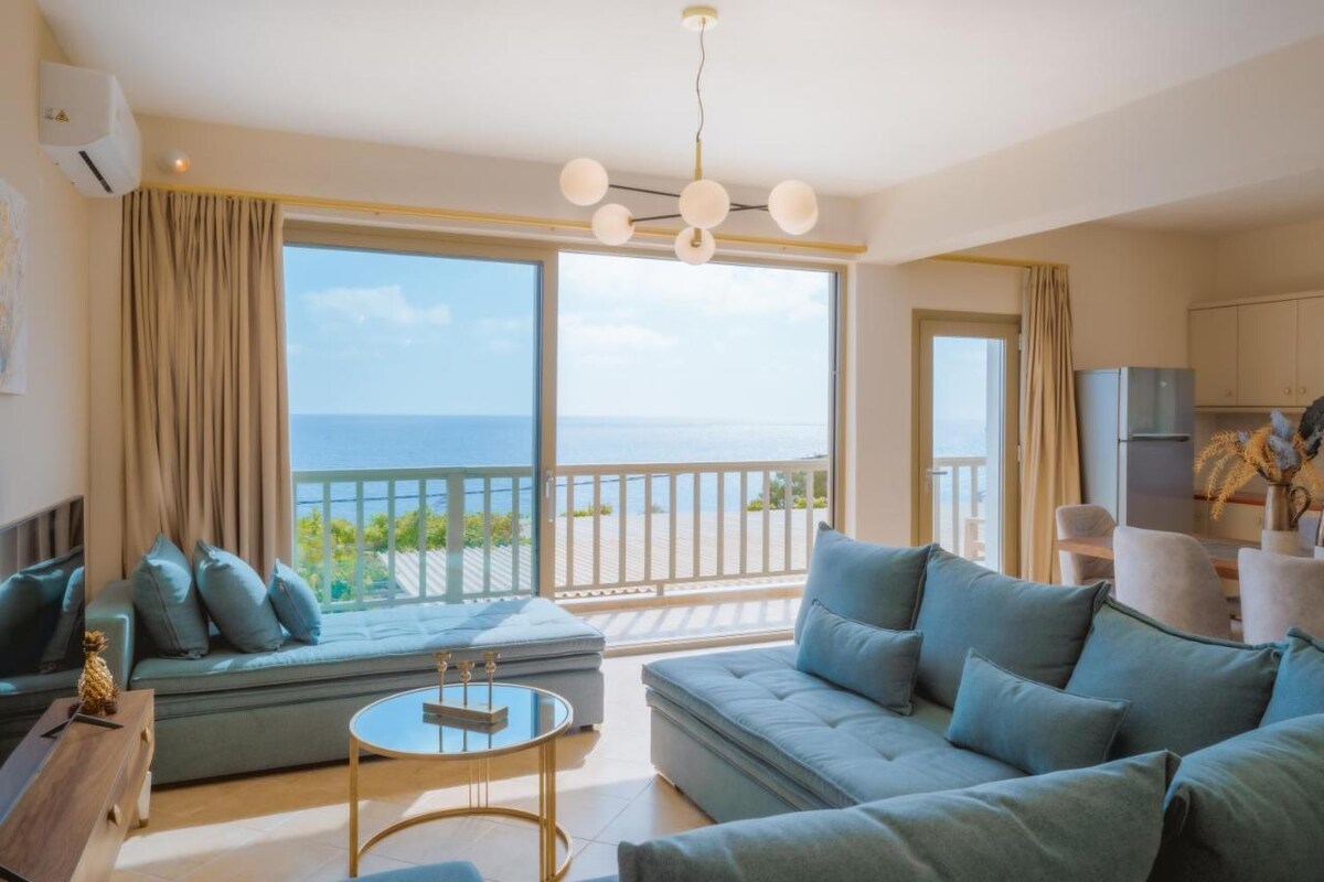 A spacious living area features large glass doors that open to a sea view. Comfortable seating arrangements include a teal sectional sofa and a modern coffee table. Soft natural light fills the room, highlighting the neutral tones and inviting décor.