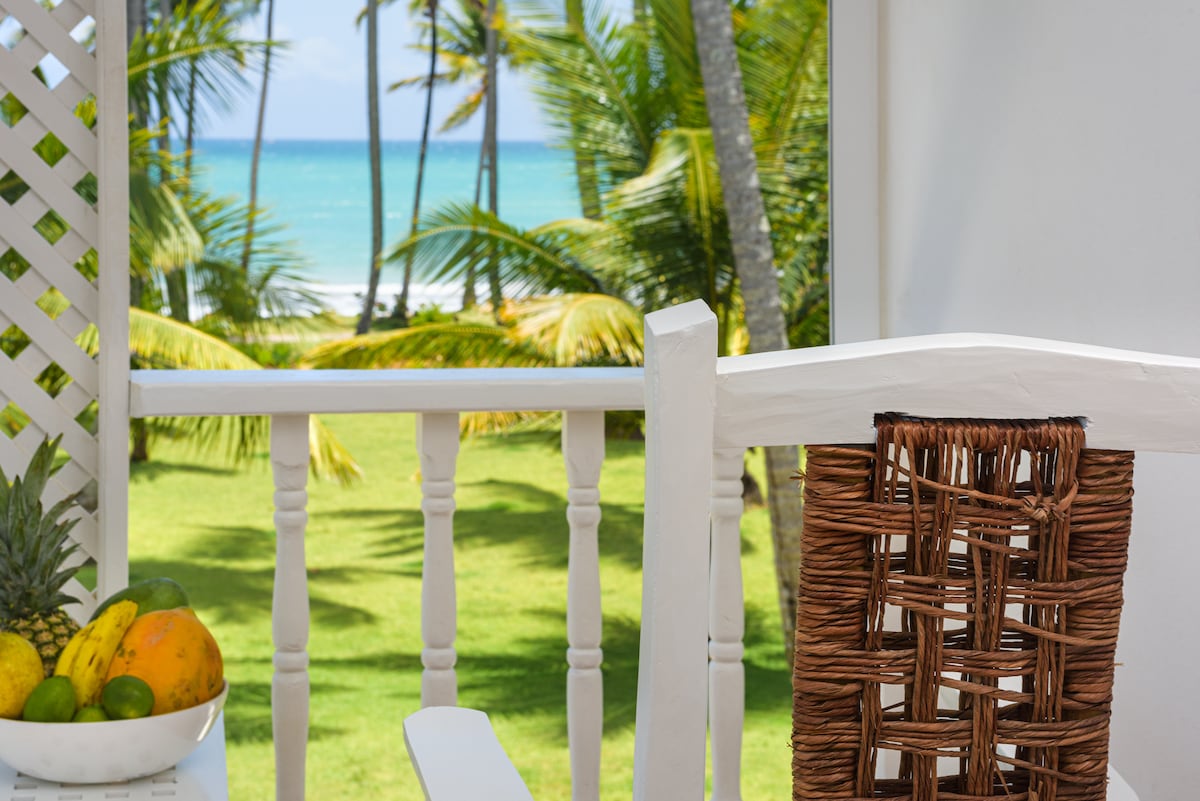 A white balcony railing frames a natural setting, featuring a view of a lush green lawn and the ocean beyond. Palm trees sway gently in the breeze. A woven chair with a textured finish complements a small table adorned with a colorful assortment of fresh fruits.