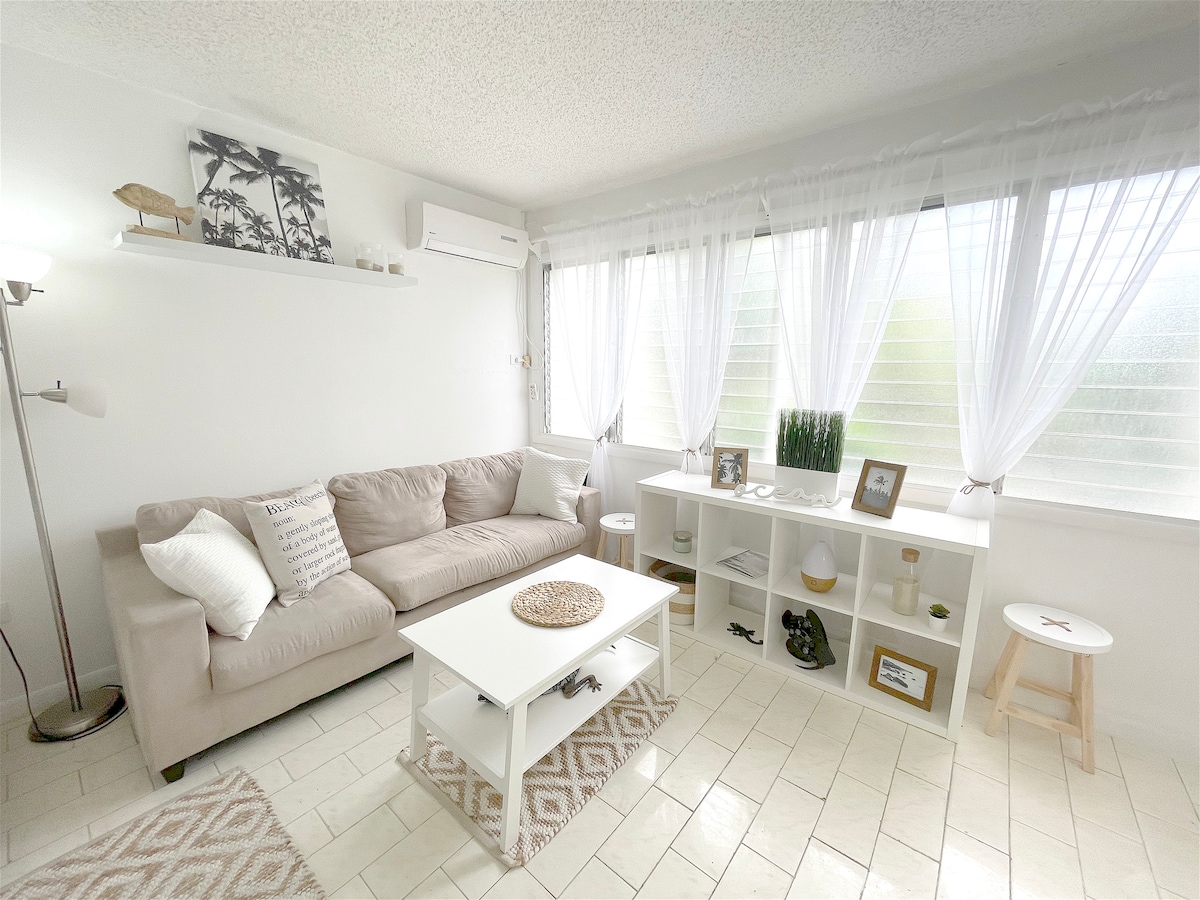A light-filled living area features a beige sofa and a white coffee table, complemented by decorative items on a shelf and side table. Sheer white curtains frame the windows, allowing natural light to create a serene ambiance. A textured area rug adds warmth to the tiled floor.