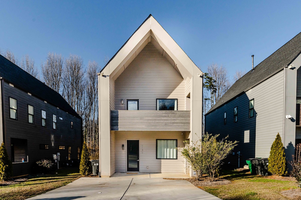 A modern Scandinavian home stands prominently with a pitched roof. The exterior features light-colored siding and a small balcony. Flanking the house are neighboring structures, with a clear driveway leading to the entrance. Green shrubs and trees provide a natural border around the property.