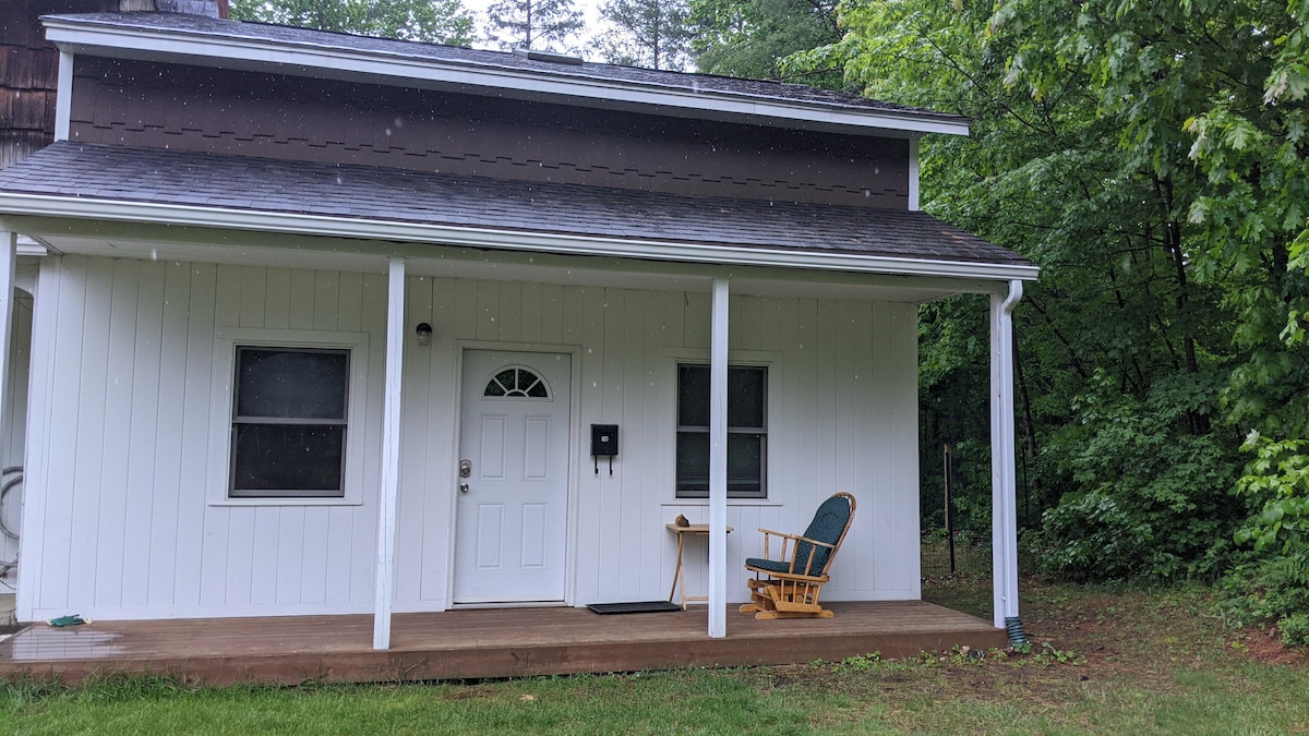 A welcoming entrance features a covered porch with a rocking chair, complemented by a small side table. The front door is framed by two windows, and a mailbox is mounted beside it. Lush greenery surrounds the exterior, enhancing the peaceful environment.