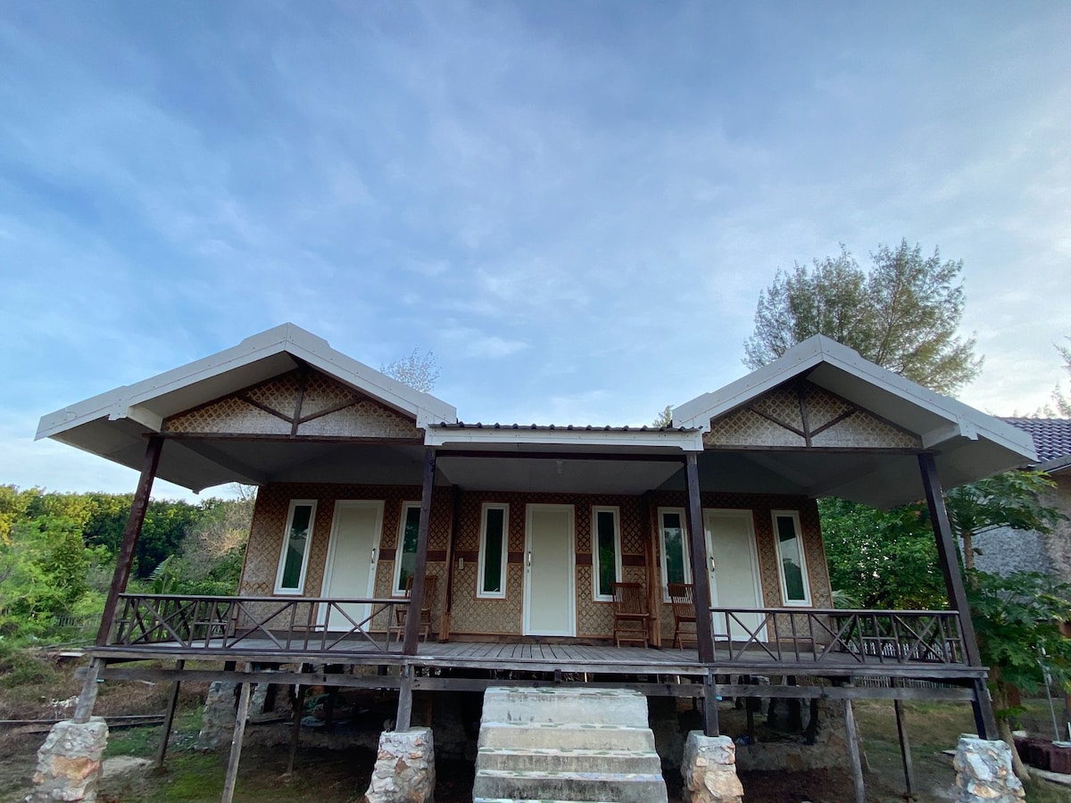 A modern bungalow with a traditional design is situated on stilts, surrounded by greenery. The structure features a covered porch with seating and has multiple doors that provide access to the interior, blending seamlessly with the natural environment.