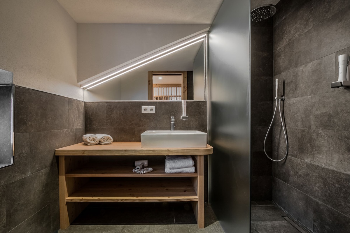 A modern bathroom features a wooden vanity with a sink, complemented by neatly arranged towels. The spacious shower area includes a sleek wall-mounted showerhead, with a frosted glass partition ensuring privacy. Soft lighting enhances the contemporary design, creating a clean and welcoming space.
