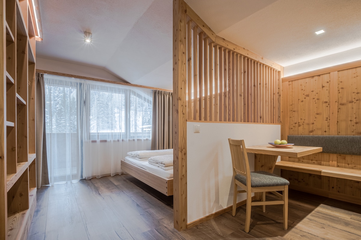 A warm, wooden interior features a spacious bed with neatly arranged bedding. A dining area includes a compact table with a fruit bowl, adjacent to a bench. Large windows are dressed with sheer curtains, allowing natural light to brighten the space.