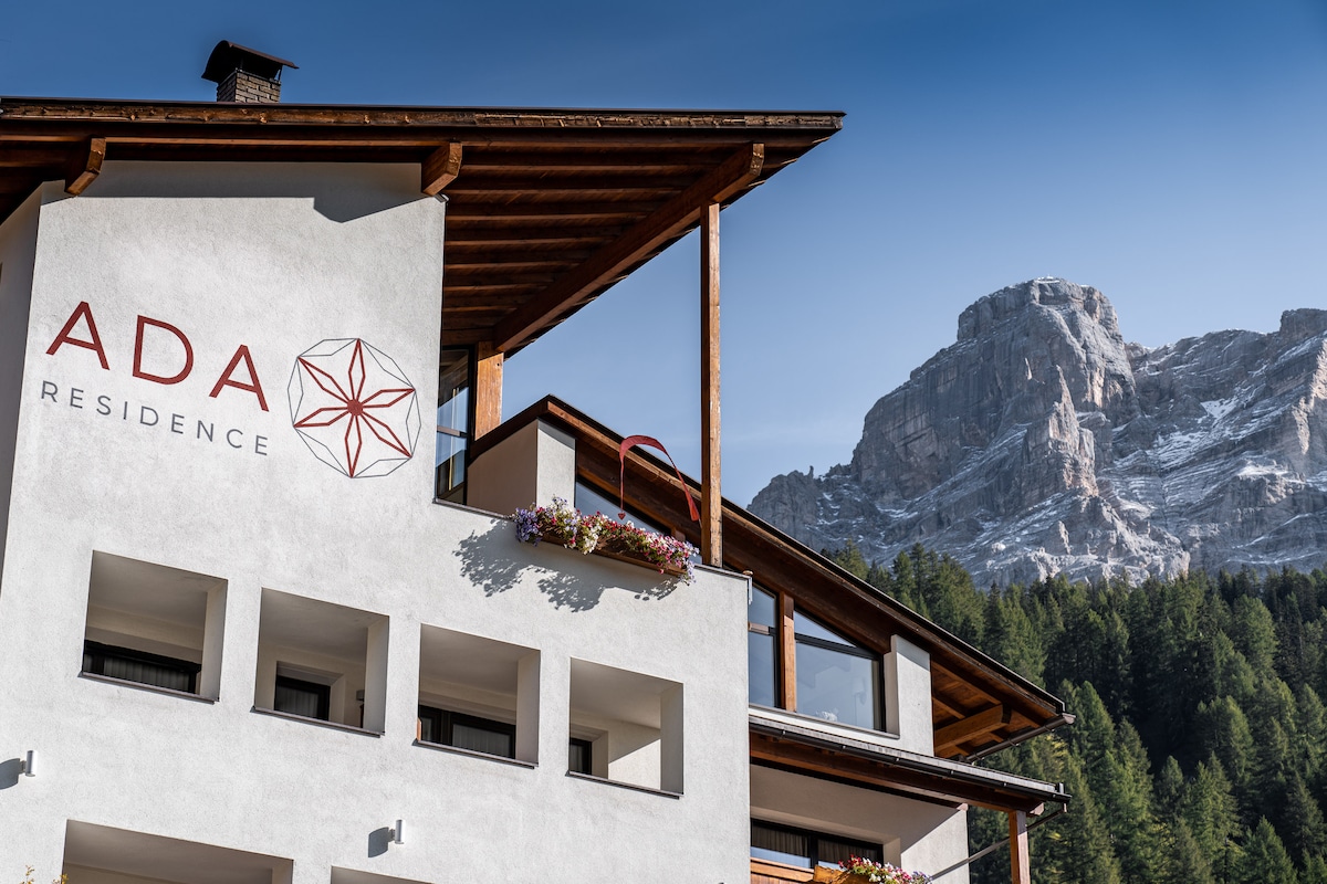 The building's exterior is showcased against a backdrop of majestic mountains. The prominent logo of 'ADA Residence' is displayed on the façade. Flower boxes can be seen at the windows, enhancing the overall rustic charm of the residence.
