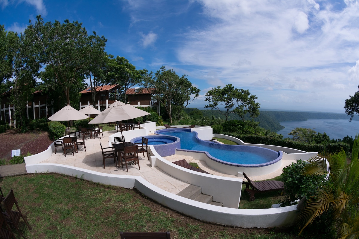 An expansive outdoor area features a large swimming pool in a unique shape, surrounded by lounge chairs and dining tables under umbrellas. Lush greenery provides a serene backdrop, while the panoramic views of Laguna de Apoyo and the sky highlight the natural beauty of the location.