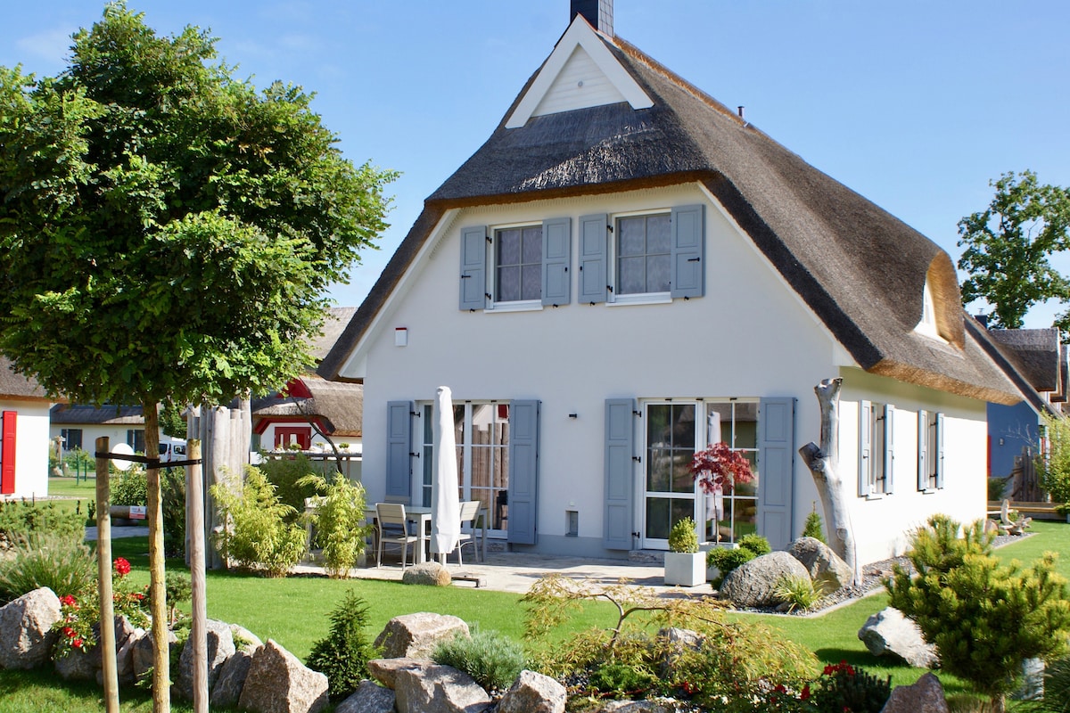 The holiday home features a traditional thatched roof and a bright exterior, set amidst neatly landscaped gardens. Several large windows provide natural light, and outdoor seating is positioned on a stone patio. Decorative rocks and trees enhance the welcoming outdoor space.