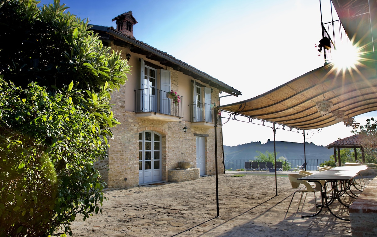 The image captures a charming stone house in a serene setting, featuring a spacious outdoor area with a large covered terrace. Sunlight filters through the canopy, illuminating nearby patio furniture, while the house's windows display floral accents against a backdrop of rolling hills.