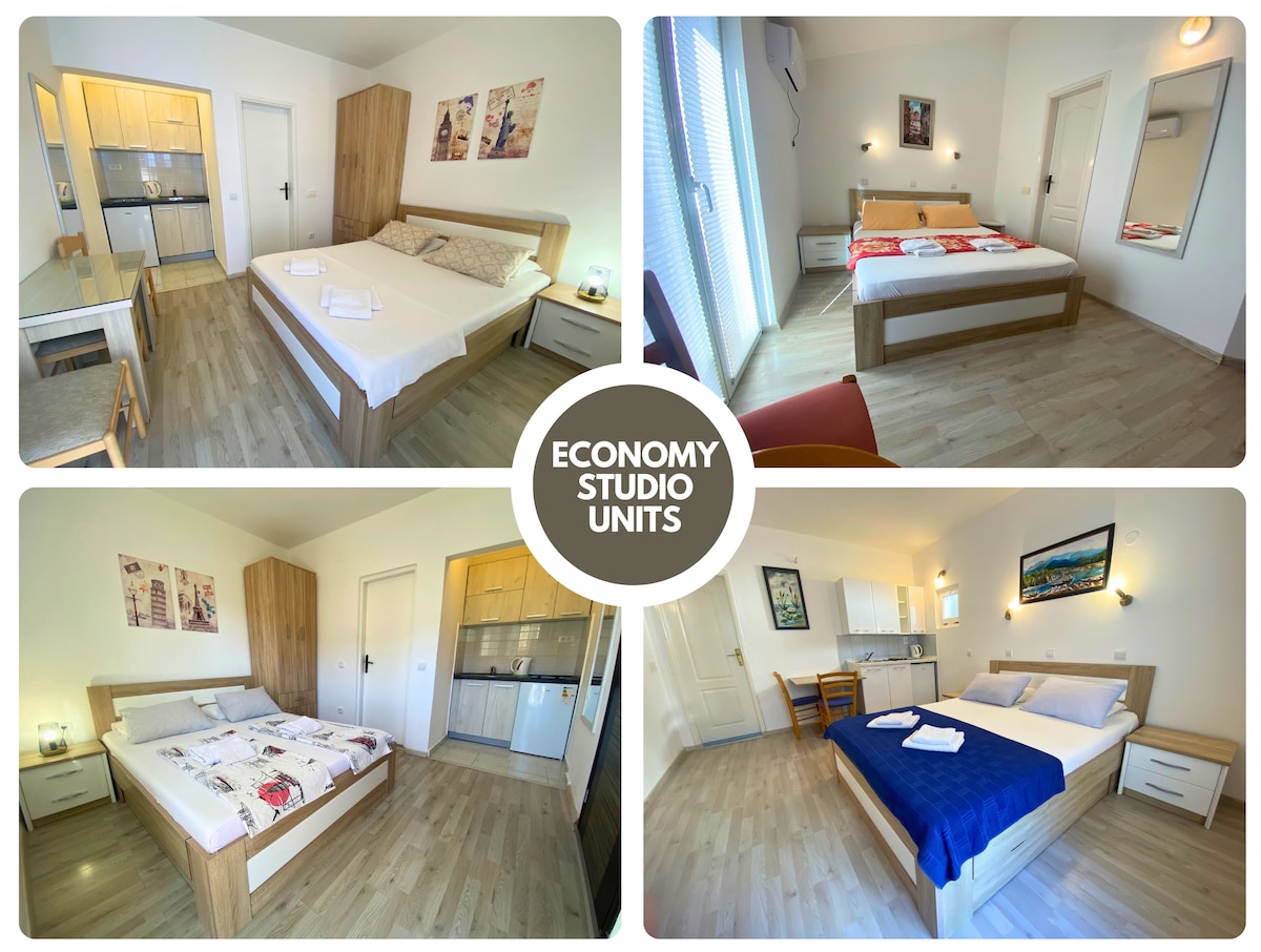 Four economy studio units are displayed, each featuring a spacious bed with varied bedding. A kitchenette is visible in the background, along with a small dining area. The studios are well-lit, showcasing neutral-colored furnishings and wall decor that enhances the inviting atmosphere.