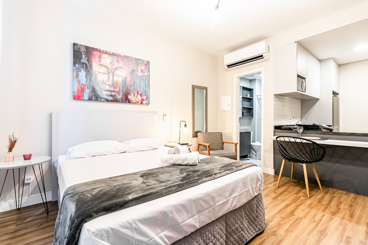 A studio room features a spacious bed with a folded blanket and decorative pillows. A small dining area with modern chairs is provided, along with a compact kitchen in the background. Natural light illuminates the space, enhancing the clean, neutral color palette.