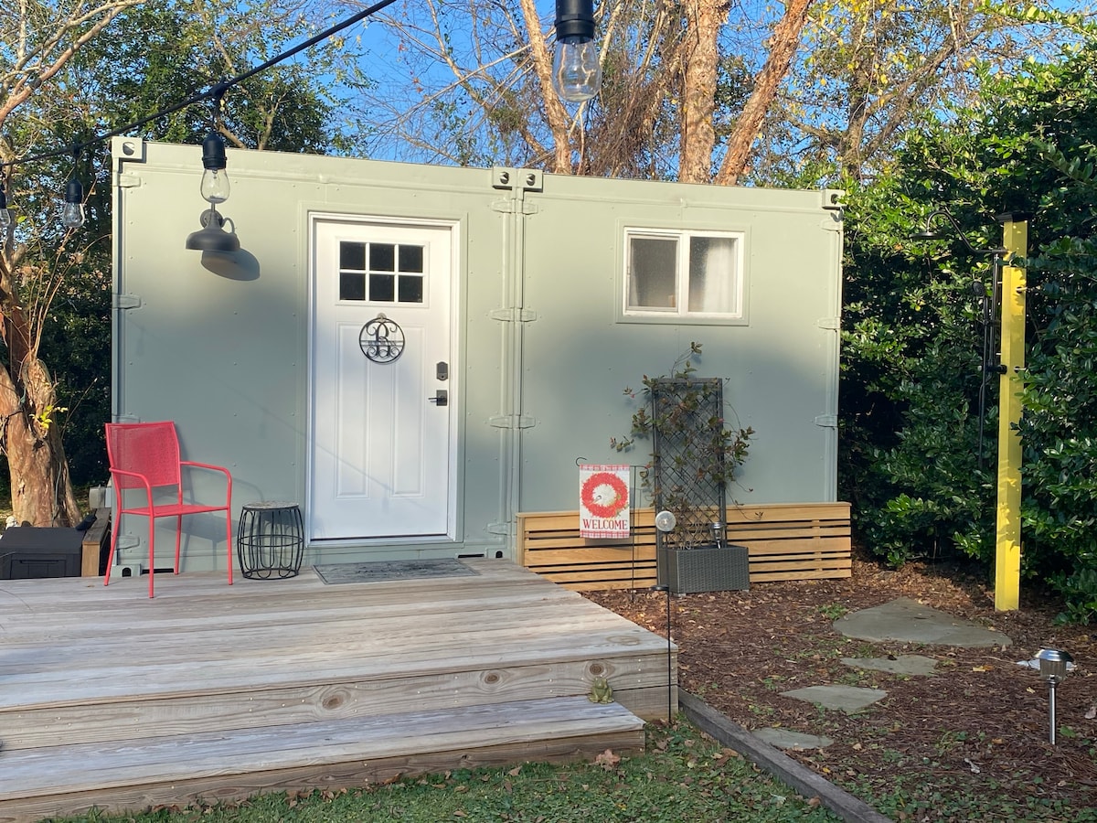 The exterior of the container home is displayed, featuring a welcoming front door framed by greenery. A deck is present, with red chairs and lighting overhead. A small sign stands next to the entrance, indicating a sense of hospitality.