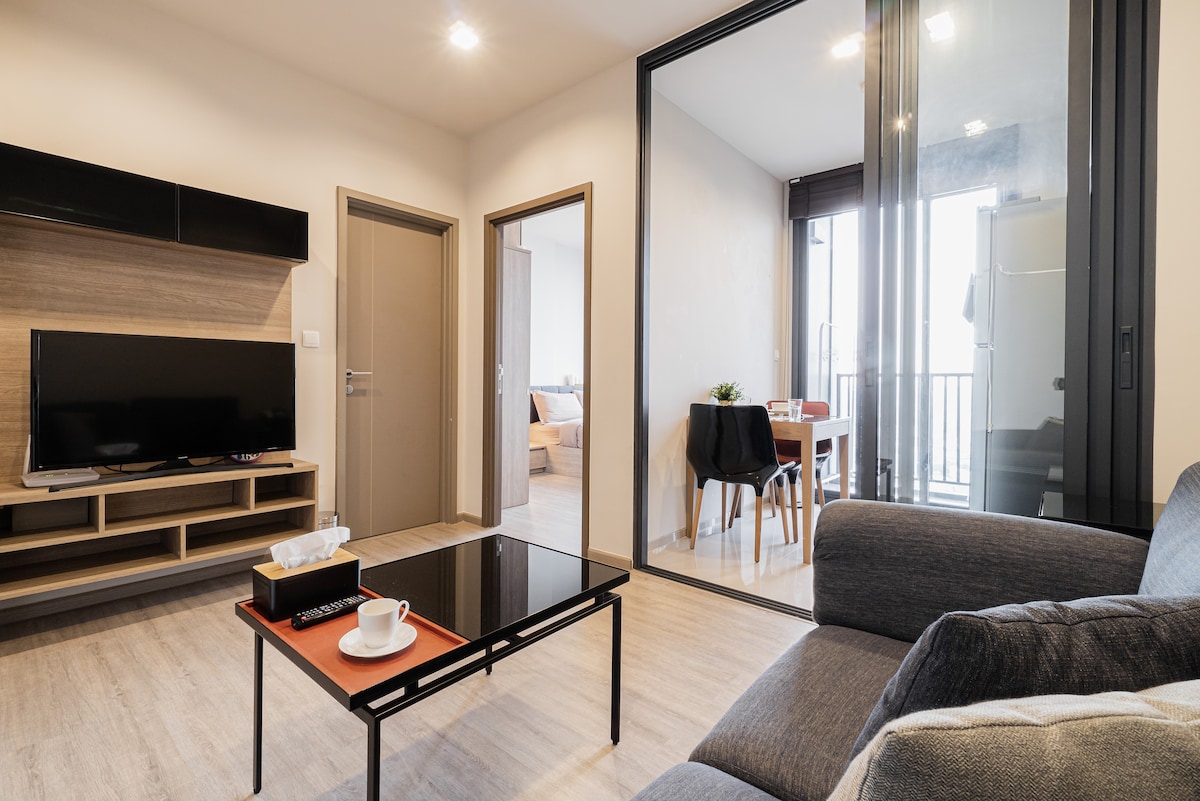 The living area features a comfortable sofa facing a mounted television. A glass door leads to a balcony that offers exterior views, while a dining area is visible in the background, showcasing a table set for four. Natural light fills the space.