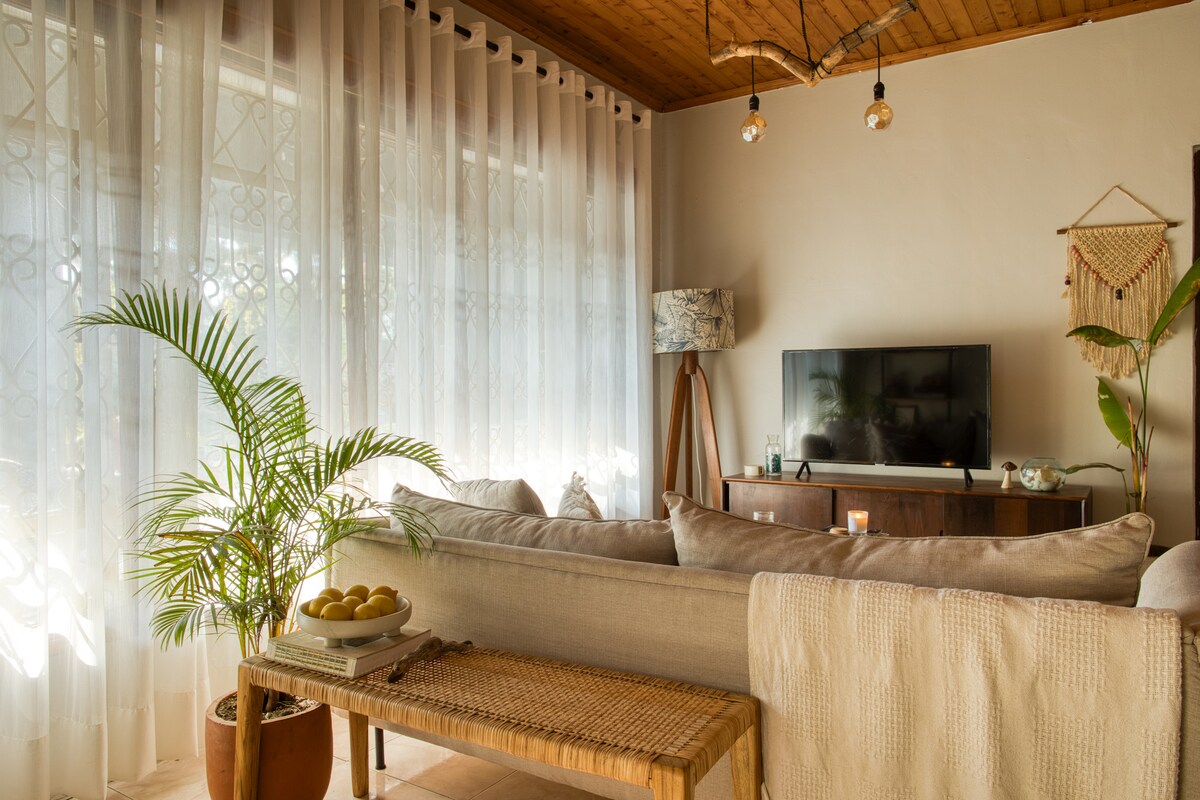 A comfortable living room is presented with a light, airy feel. Sheer white curtains allow natural light to diffuse softly across the space. A cozy sofa is paired with a wooden coffee table, which holds a bowl of fruit. A TV is visible, with decorative plants adding greenery.