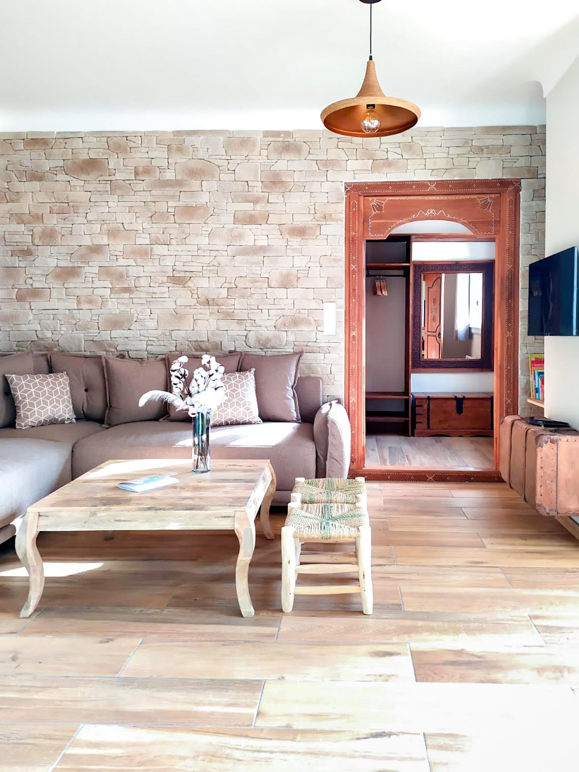 A cozy living area is highlighted by a stone-patterned wall and a comfortable sectional sofa. A wooden coffee table sits in front, accompanied by a small, woven stool. To the right, an archway leads to another room, while a television is mounted on the opposite wall.