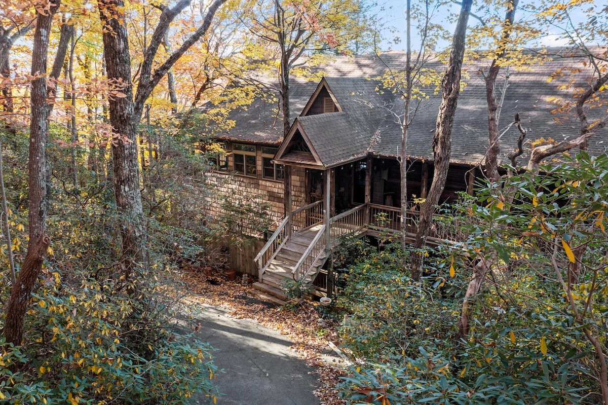 The exterior of a mountain lodge is framed by colorful autumn foliage. A wooden walkway leads to the entrance, bordered by greenery. The shingled structure blends into the natural surroundings, showcasing a sense of tranquility and connection with nature.
