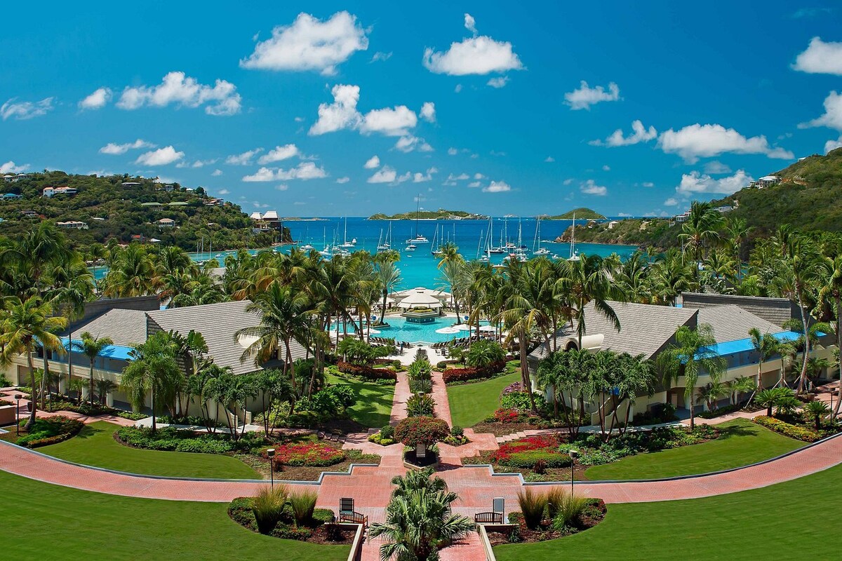 A panoramic view of the resort highlights vibrant gardens lined with tropical plants and palm trees. In the background, turquoise waters and a harbor with anchored yachts reflect the clear blue sky, creating a serene coastal atmosphere.