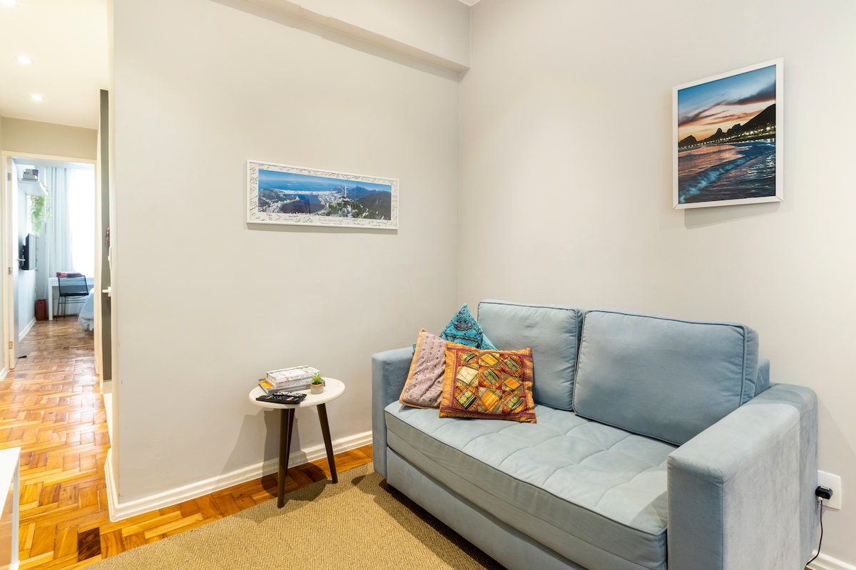 A light and airy living area features a blue sofa with decorative pillows, complemented by a small round table. A piece of framed artwork hangs on the wall, depicting a scenic view. Natural light enhances the warm wood flooring and muted walls.